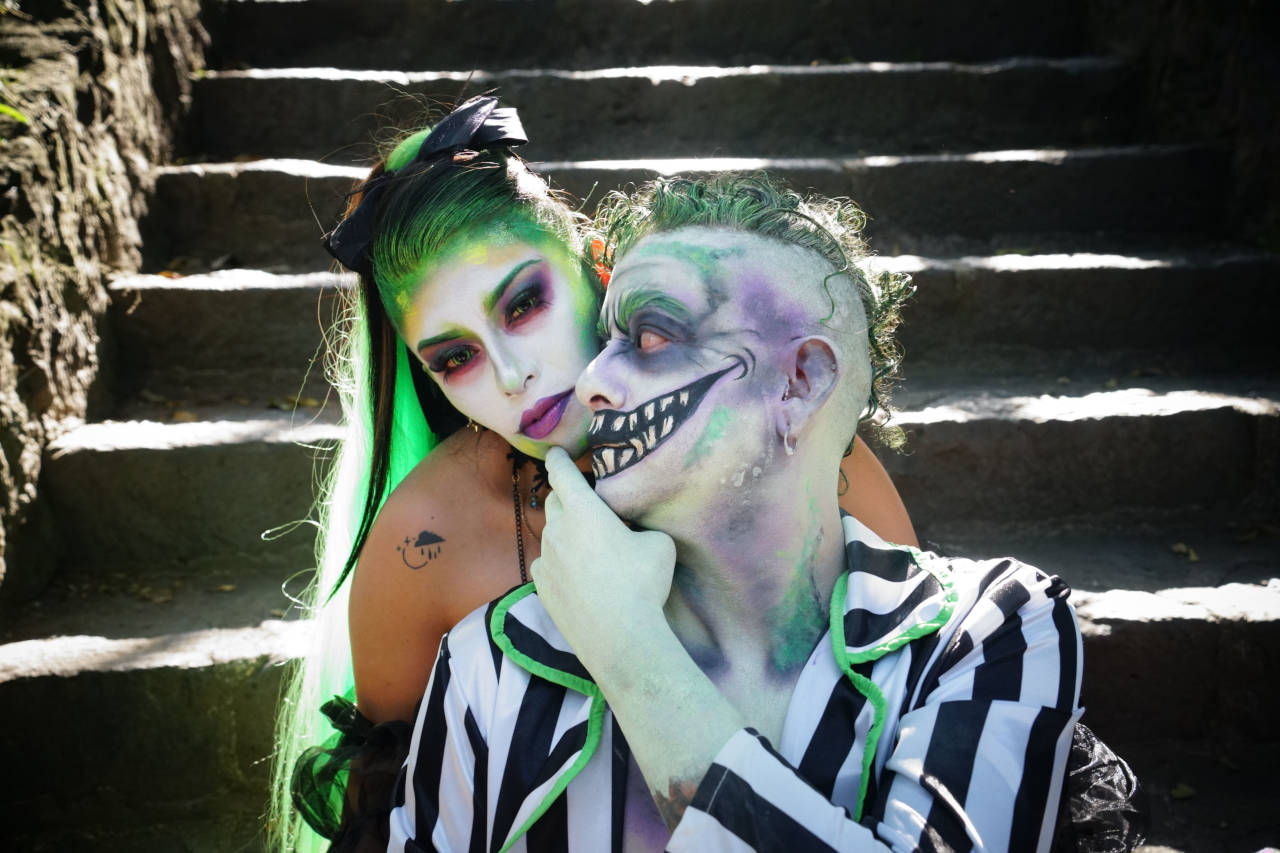 Two people dressed in Beetlejuice-inspired Halloween costumes with green hair, dramatic makeup, and black-and-white striped clothing pose together on outdoor stone steps.
