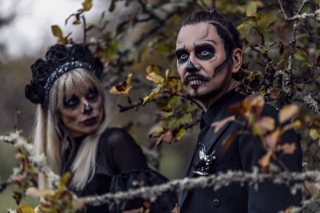 Two people in gothic Halloween costumes with dramatic skull-inspired face makeup and black clothing pose among autumn trees and branches.