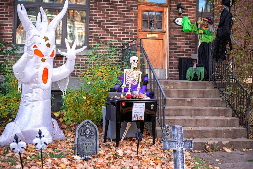 Halloween-decorated house with a giant inflatable white ghost, skeleton, witch, grim reaper, and spooky tombstones in a front yard covered with autumn leaves.