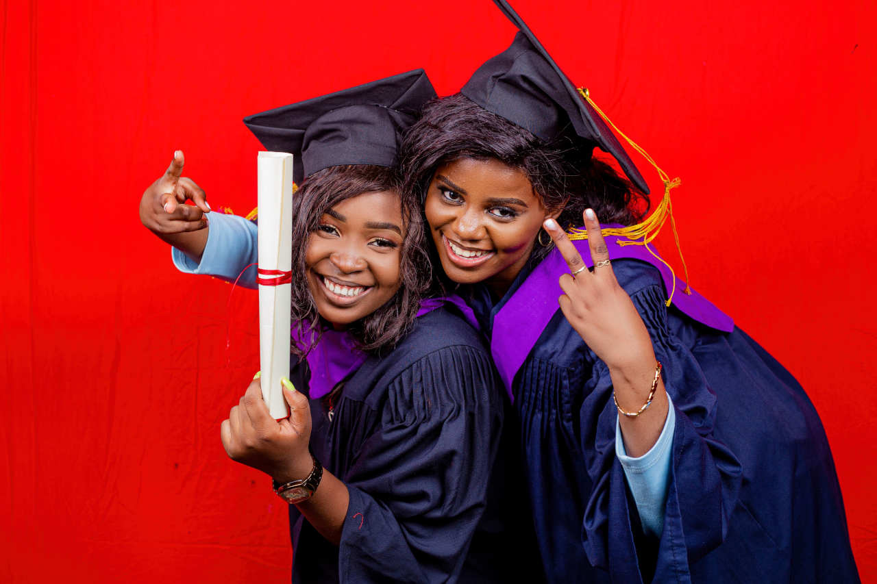 Two graduates in caps and gowns smiling and celebrating while holding a diploma against a bright red background.