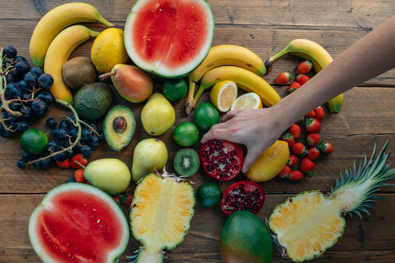 Assorted fresh fruits including watermelon, bananas, avocado, grapes, berries, citrus, pineapple and pomegranate on a wooden table.