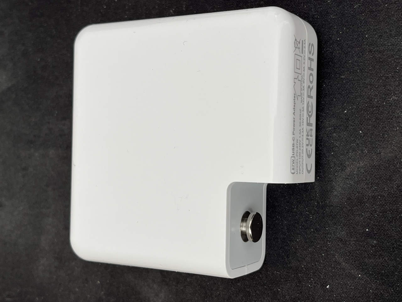 No-name 87W USB-C MacBook power adapter photographed from the side on a dark surface.