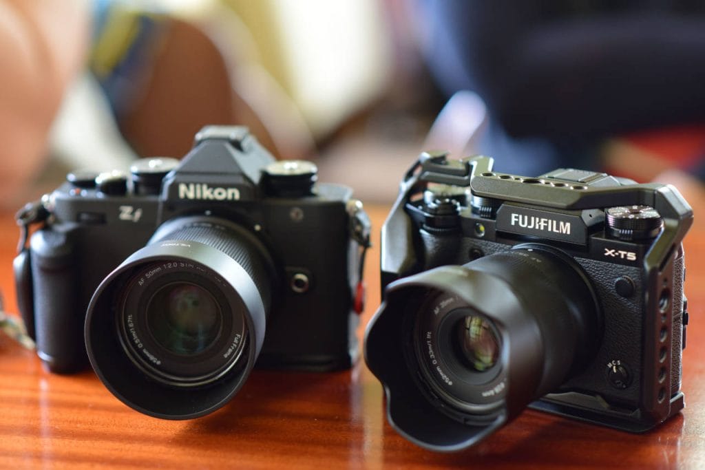 Nikon Zf and Fujifilm X-T5 mirrorless cameras with attached lenses displayed side by side on a wooden table.