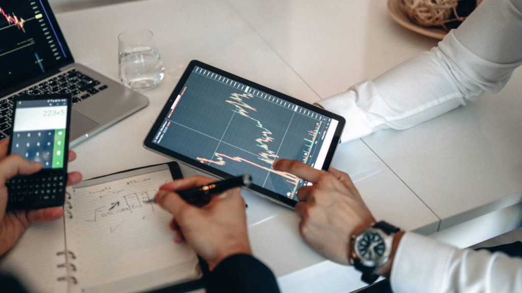 Business professionals analyzing financial market charts on a tablet and laptop while using a calculator and taking notes during an investment discussion.