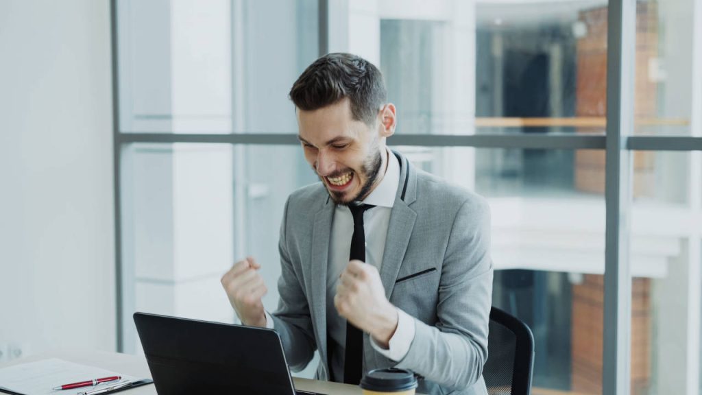 Smiling businessman in a gray suit celebrating success with clenched fists while working on a laptop in a modern office.