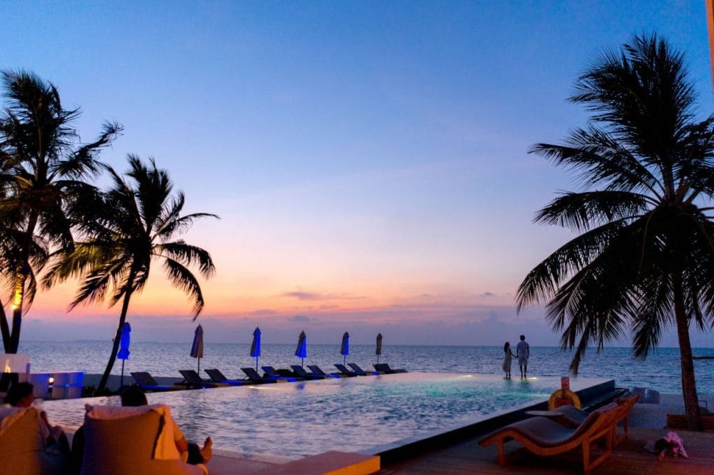 Tropical beach resort at sunset with palm trees, an infinity pool, sun loungers, and a couple standing by the ocean.