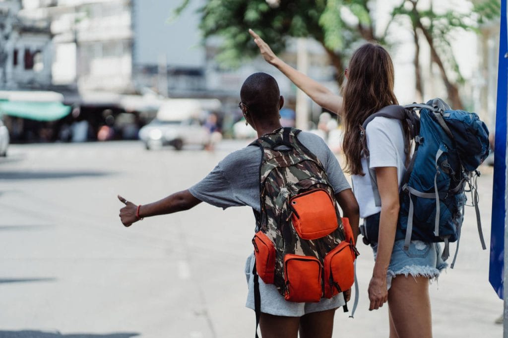Two backpackers hitchhiking on a city street, with one giving a thumbs-up and the other waving to passing vehicles.