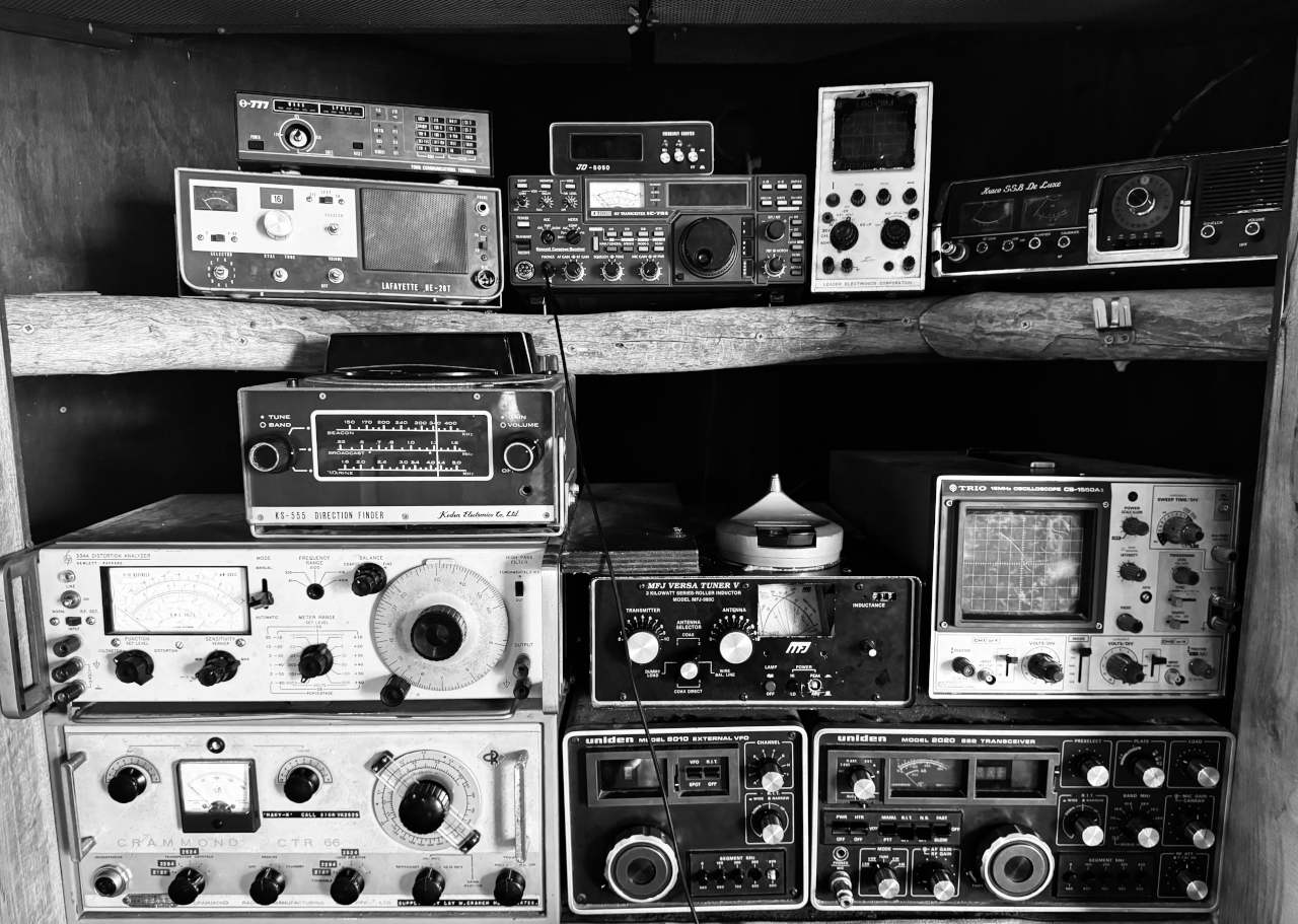 Black-and-white photograph of vintage amateur radio transceivers, receivers, antenna tuners, and electronic test equipment arranged on wooden shelves.